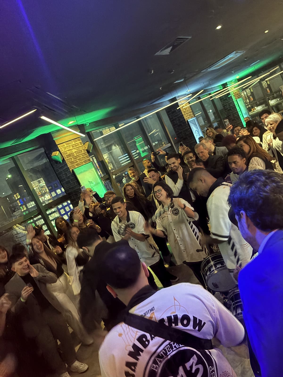 Convidados formam uma roda em torno de ritmistas de camisa branca, filmando com celulares em um salão com neon verde.