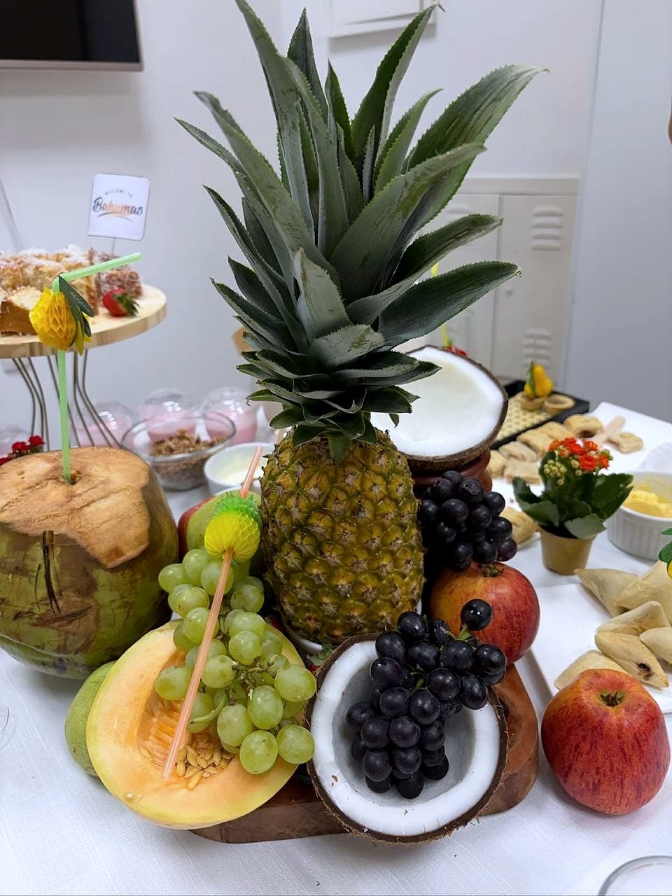 Composição de frutas com abacaxi inteiro, cocos abertos, cachos de uva, melão e maçãs sobre mesa branca.