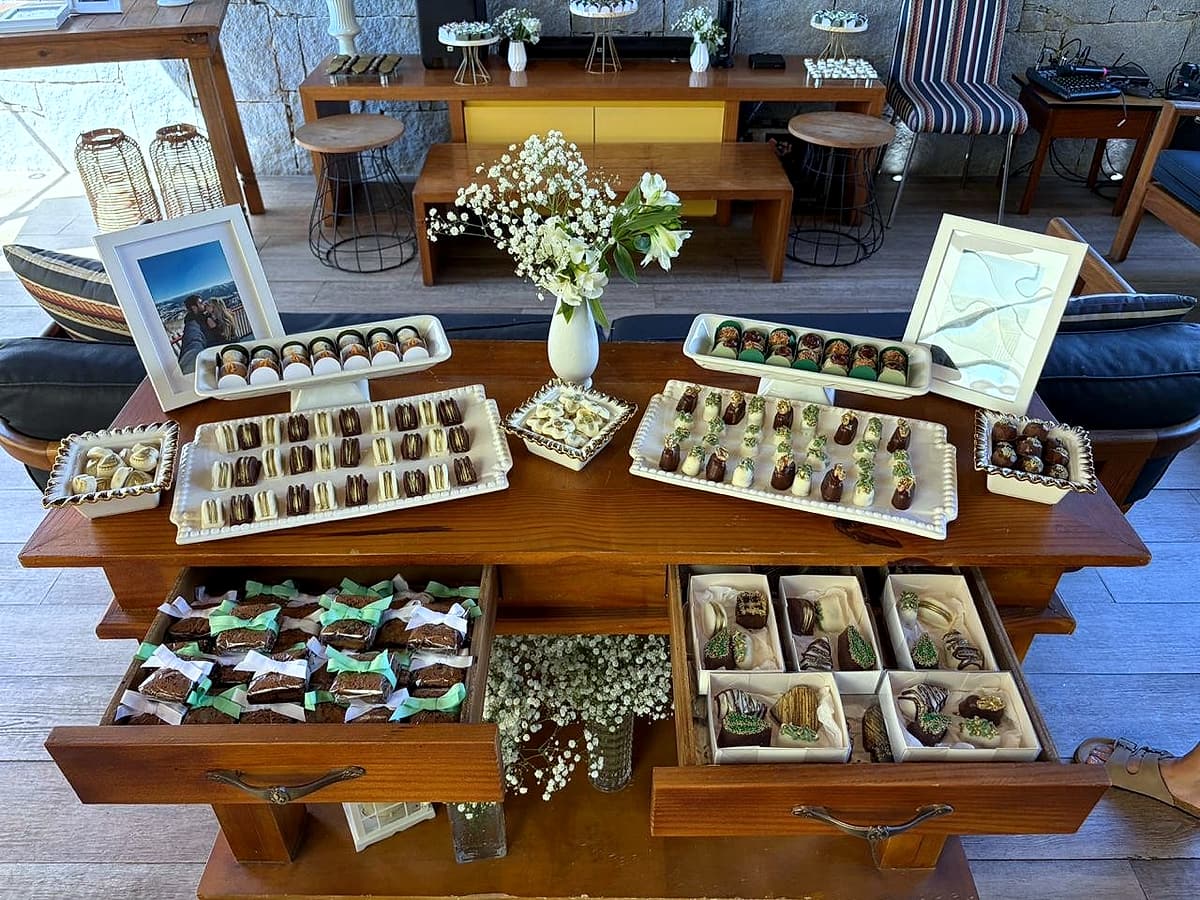 Cômoda de madeira com bandejas de doces e bombons sobre o tampo e nas gavetas abertas, com arranjo de flores brancas.