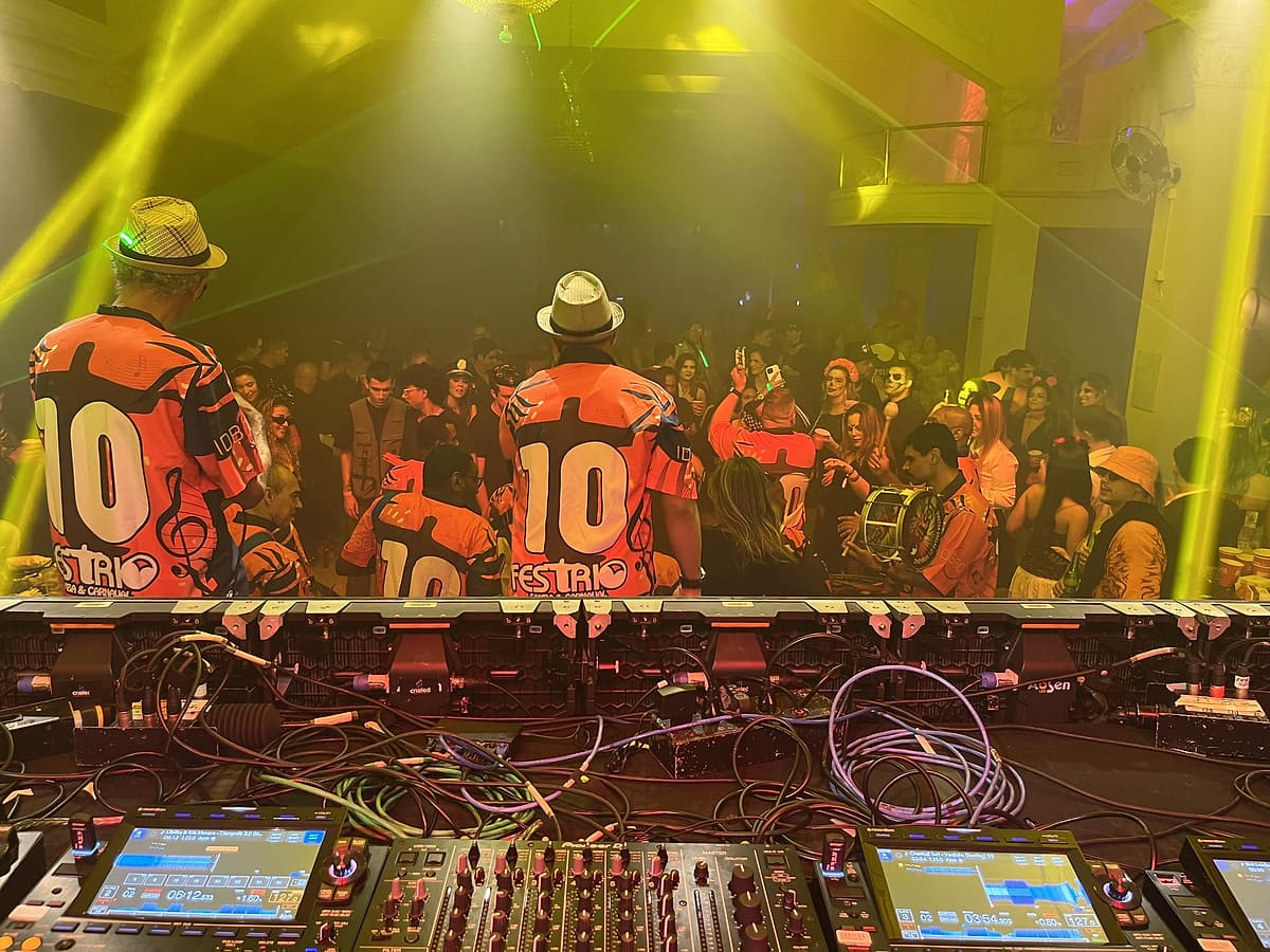 Vista da cabine de DJ com mixers em primeiro plano; ritmistas de camisa laranja com o número 10 de costas, diante da pista lotada sob lasers amarelos.