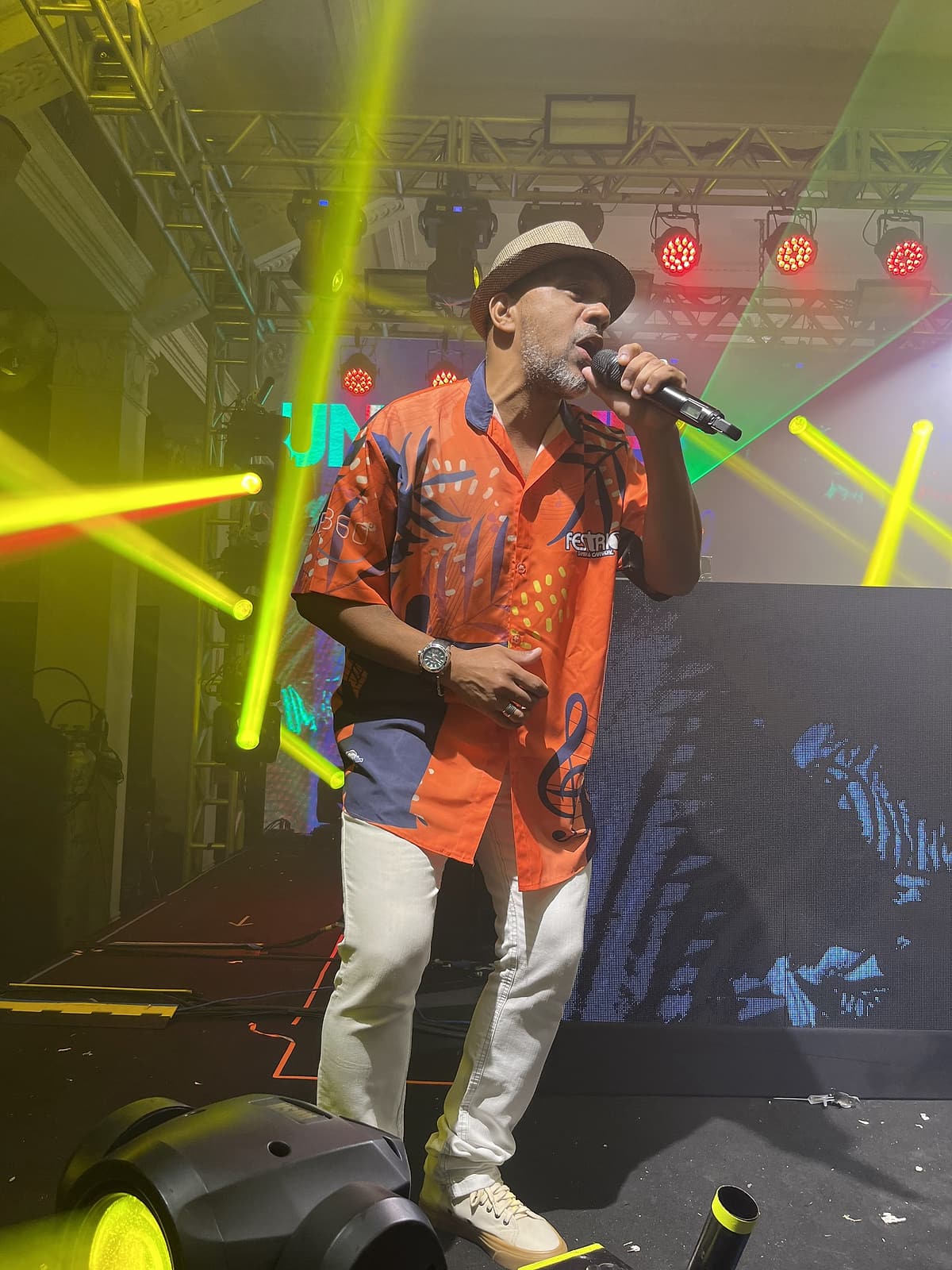 Cantor de chapéu panamá e camisa laranja canta ao microfone no palco, com feixes de laser verde-limão ao fundo.