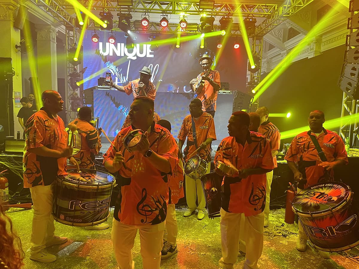 Ritmistas de camisa laranja tocam surdos e tamborins em frente a um palco com treliças, lasers verde-limão e telão de LED.