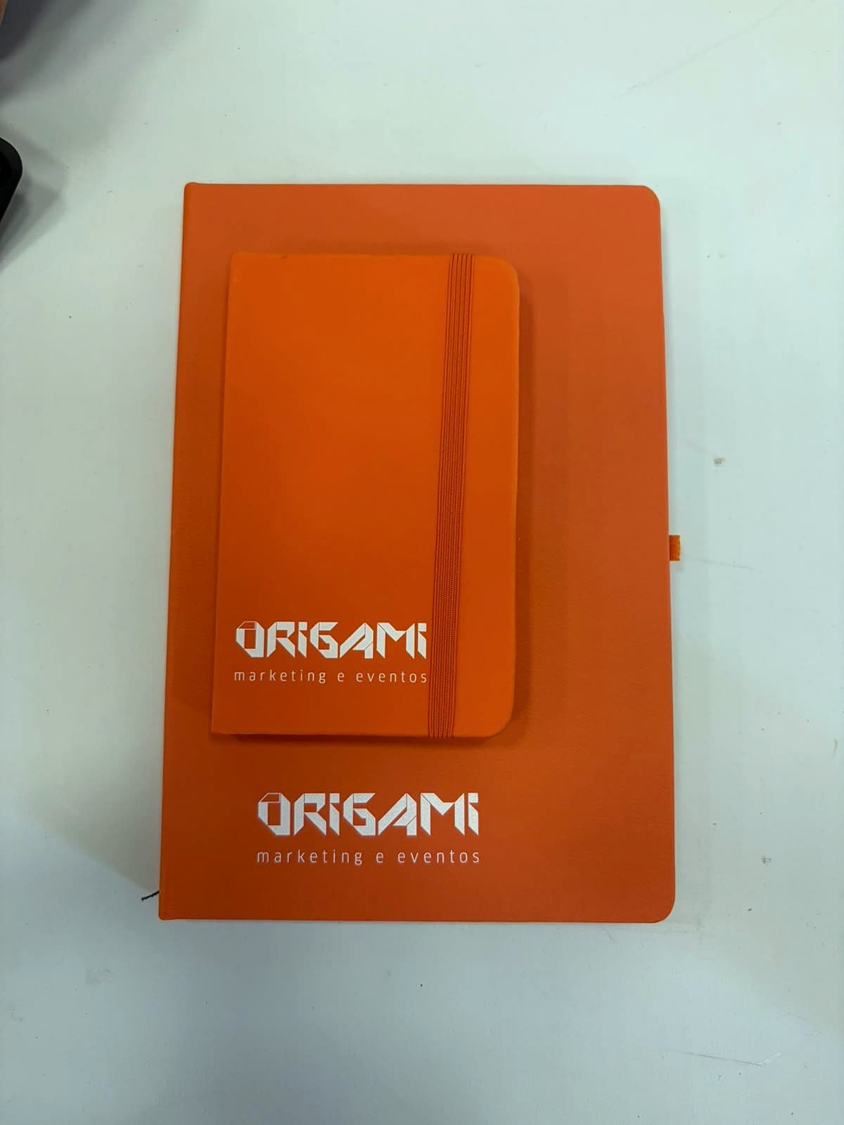 Caderno laranja grande e caderno de bolso laranja sobrepostos, ambos com o logo Origami Marketing e Eventos em branco.