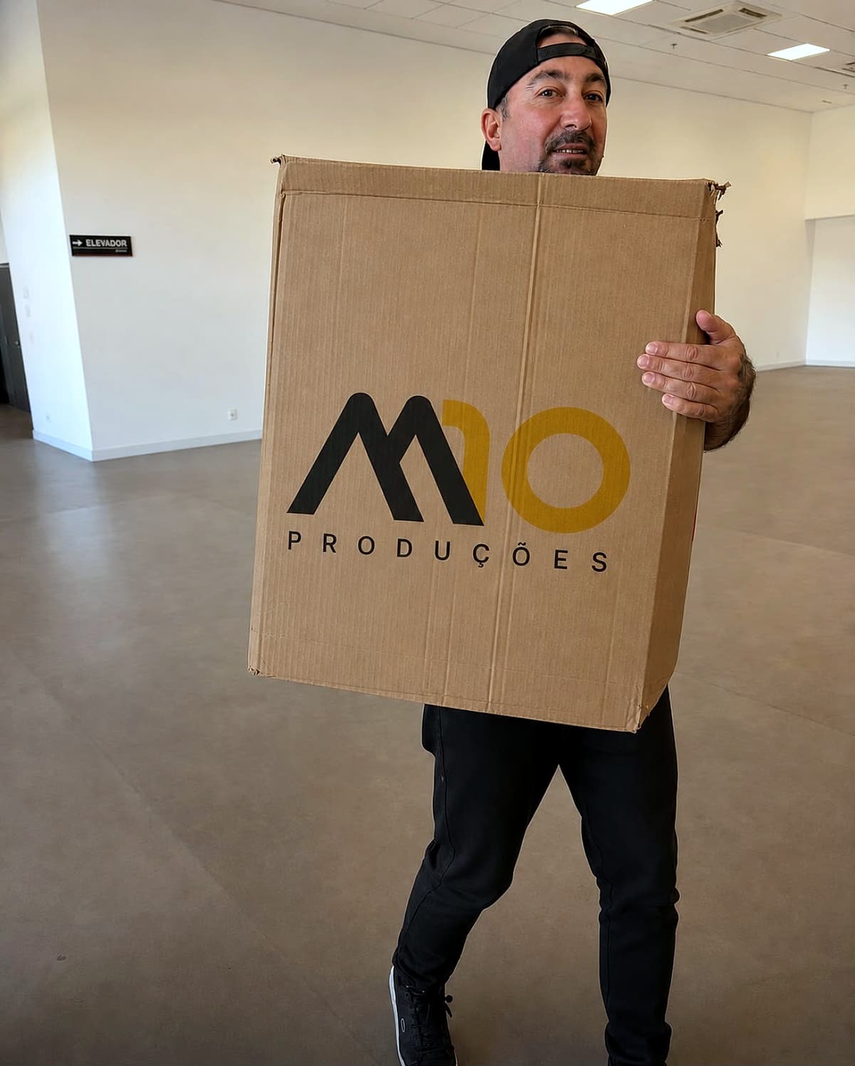 Homem de boné carregando uma grande caixa de papelão com o logo M10 Produções impresso, em um salão vazio.