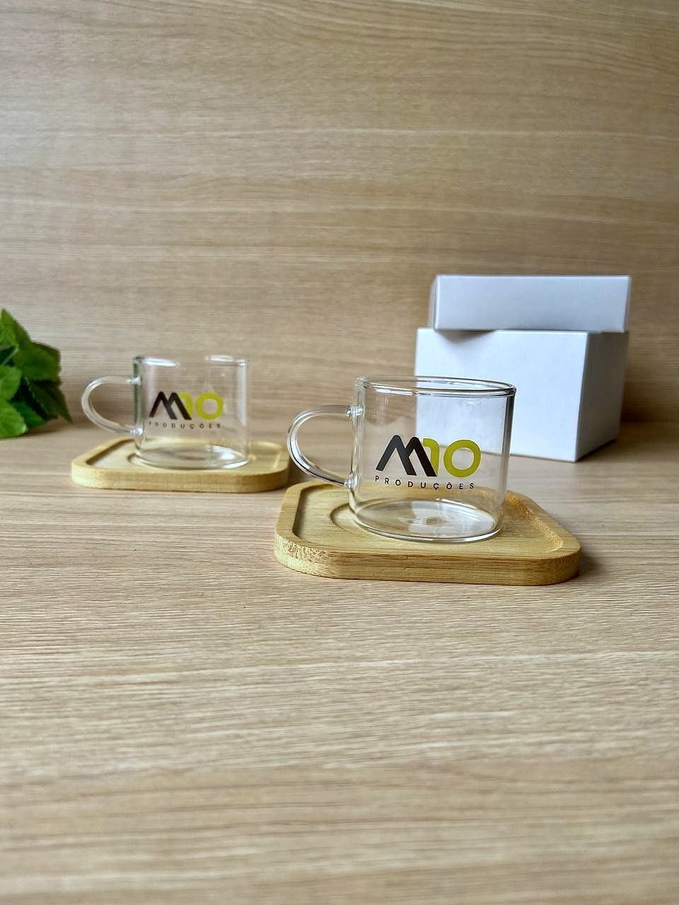 Duas xícaras de vidro transparentes com o logo M10 Produções sobre pires de bambu, com caixa branca e folhagem ao fundo em madeira clara.