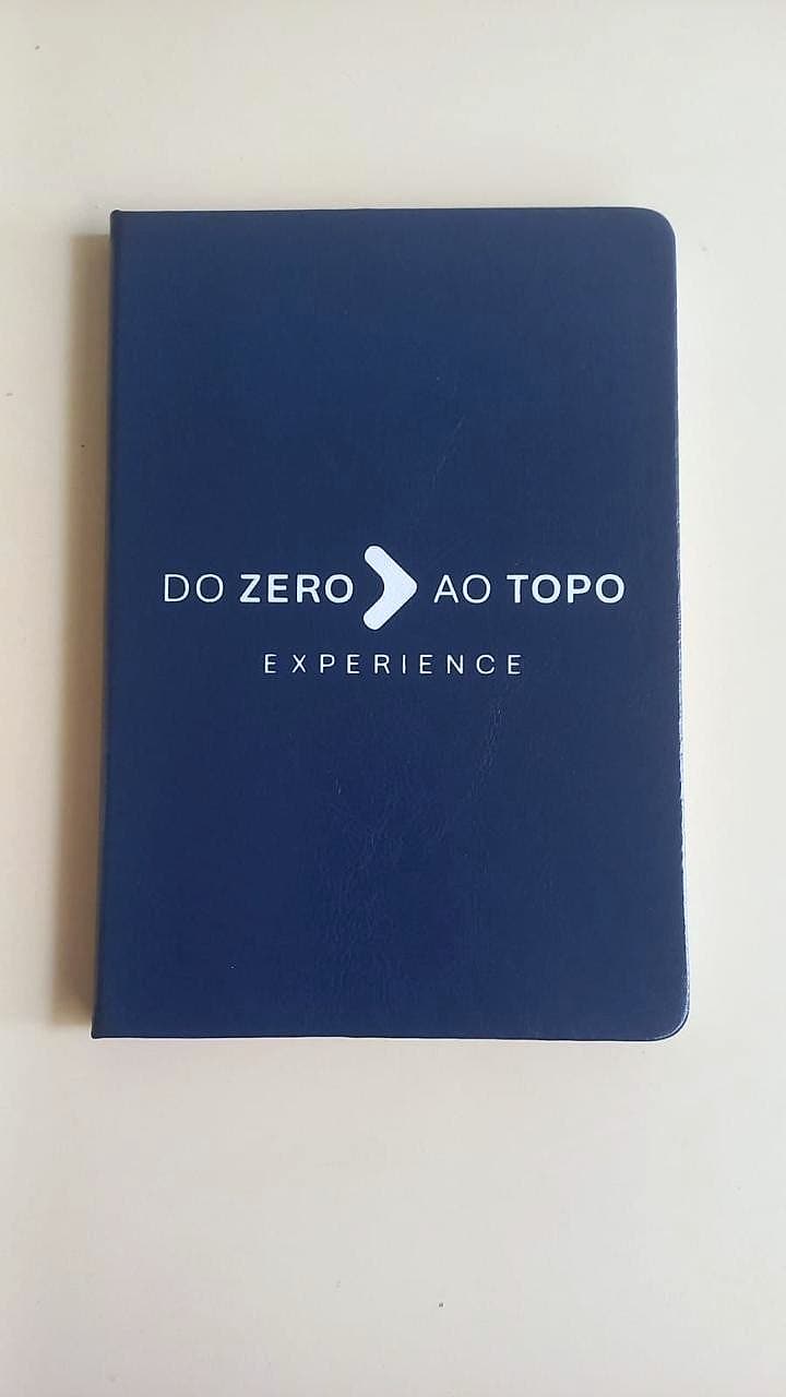 Caderno azul-marinho com a marca 'Do Zero ao Topo Experience' impressa em branco sobre fundo creme.