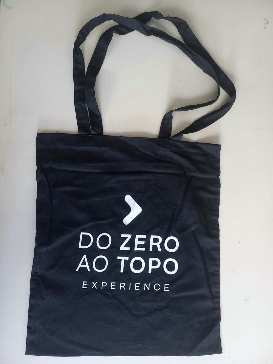 Ecobag preta de algodão com a marca 'Do Zero ao Topo Experience' em branco, estendida sobre mesa creme.