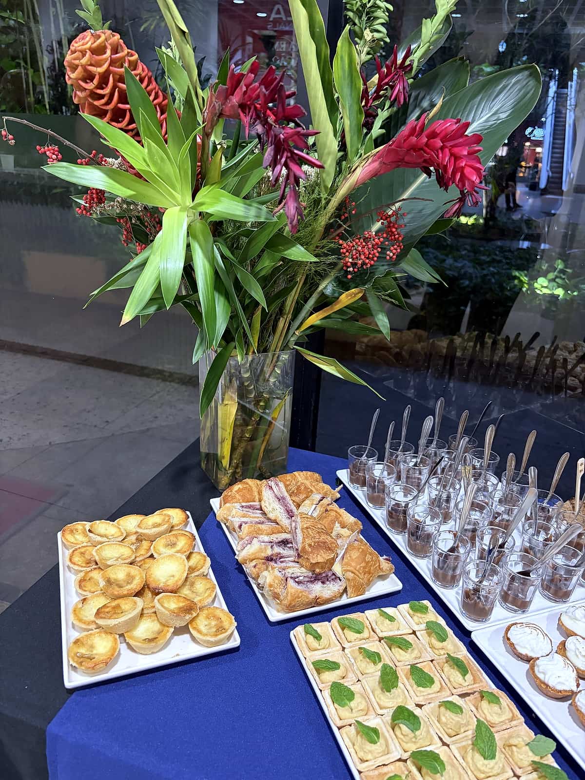 Arranjo de flores tropicais vermelhas sobre mesa azul com bandejas de empadinhas, folhados, canapés e copinhos de doce.