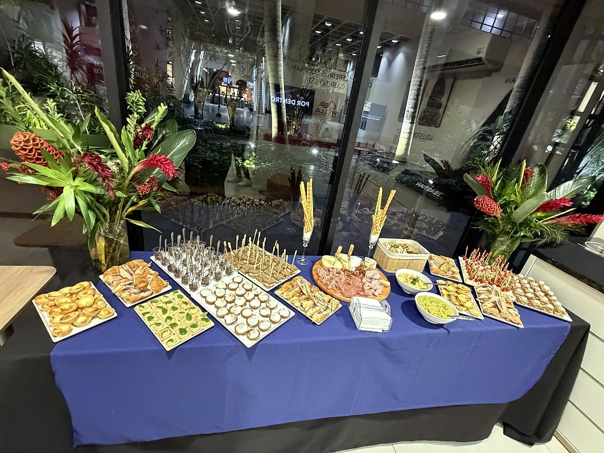 Mesa com toalha azul coberta de bandejas de salgados e doces, com arranjos de flores tropicais nas duas pontas.
