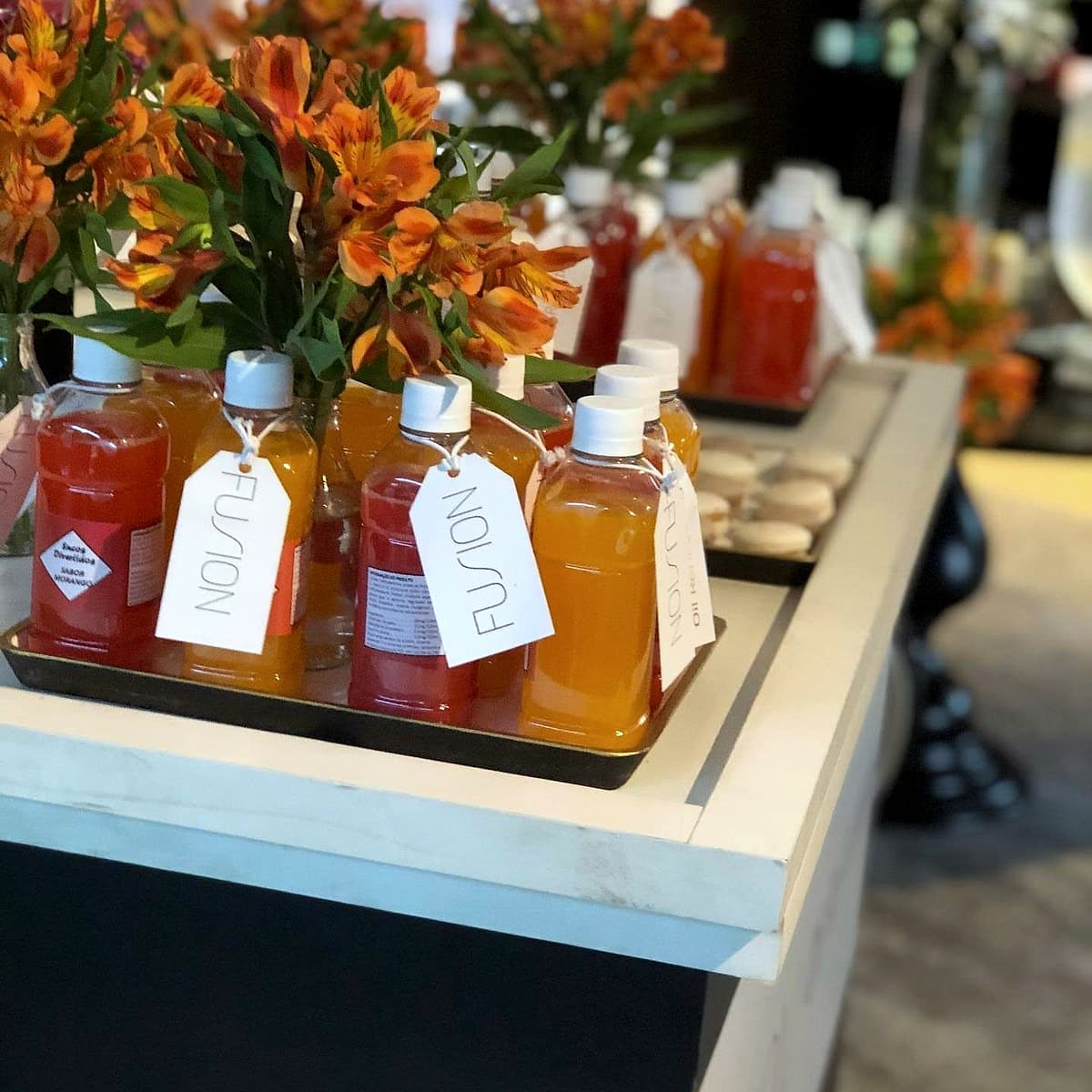 Garrafinhas de suco vermelho e laranja com etiquetas penduradas escritas 'FUSION', ao lado de flores laranja.