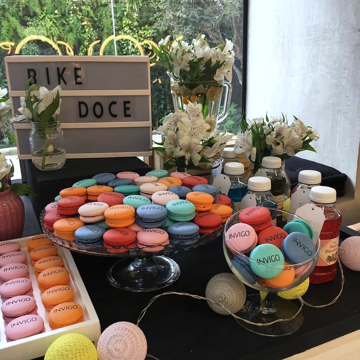 Letreiro 'BIKE DOCE' sobre mesa com dezenas de macarons coloridos estampados INVIGO, flores brancas e sucos.