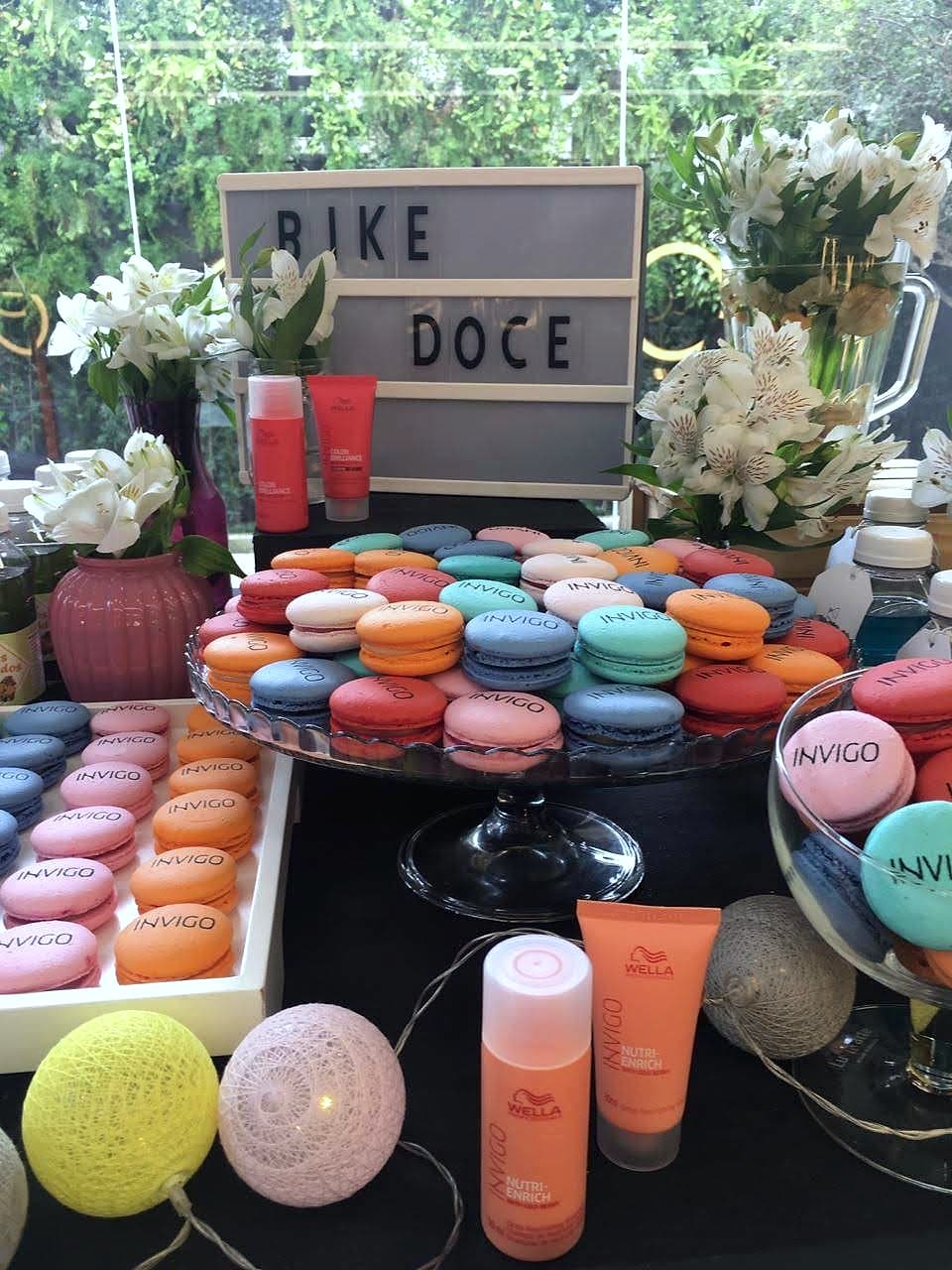 Mesa com letreiro 'BIKE DOCE', prato de macarons coloridos, luminárias de fio e frascos laranja Wella INVIGO.