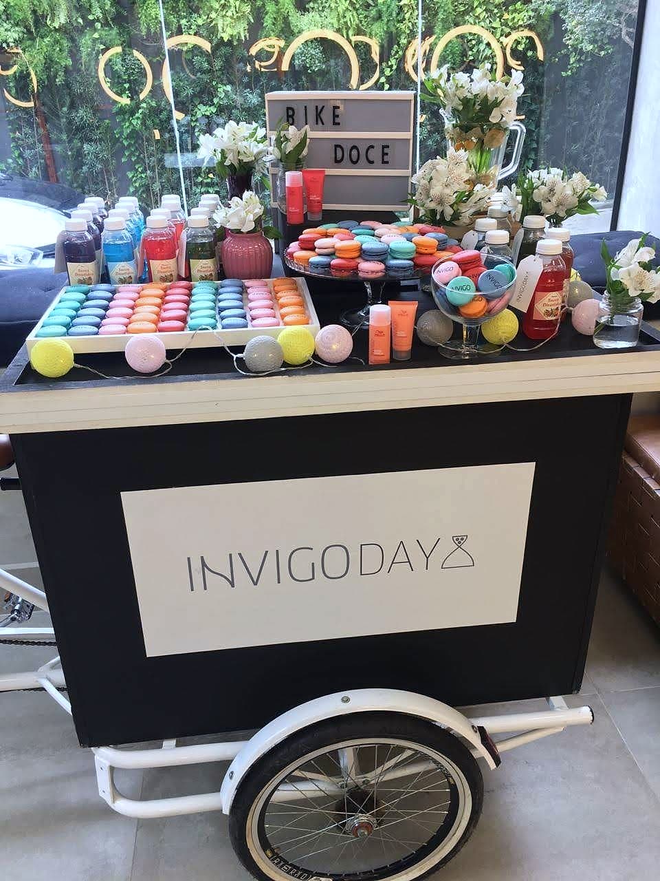 Triciclo preto com painel 'INVIGO DAY', tampo com macarons coloridos, sucos e flores brancas.