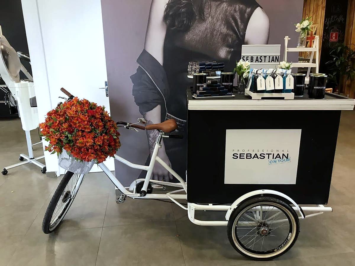 Triciclo branco com baú preto e placa 'Sebastian Professional On Tour', cesto dianteiro com flores laranja.