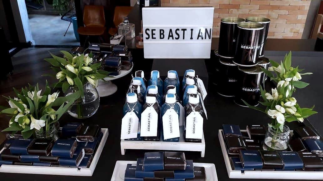 Letreiro 'SEBASTIAN' sobre mesa preta com caixinhas azuis de produto, garrafinhas com tags e arranjos de flores brancas.