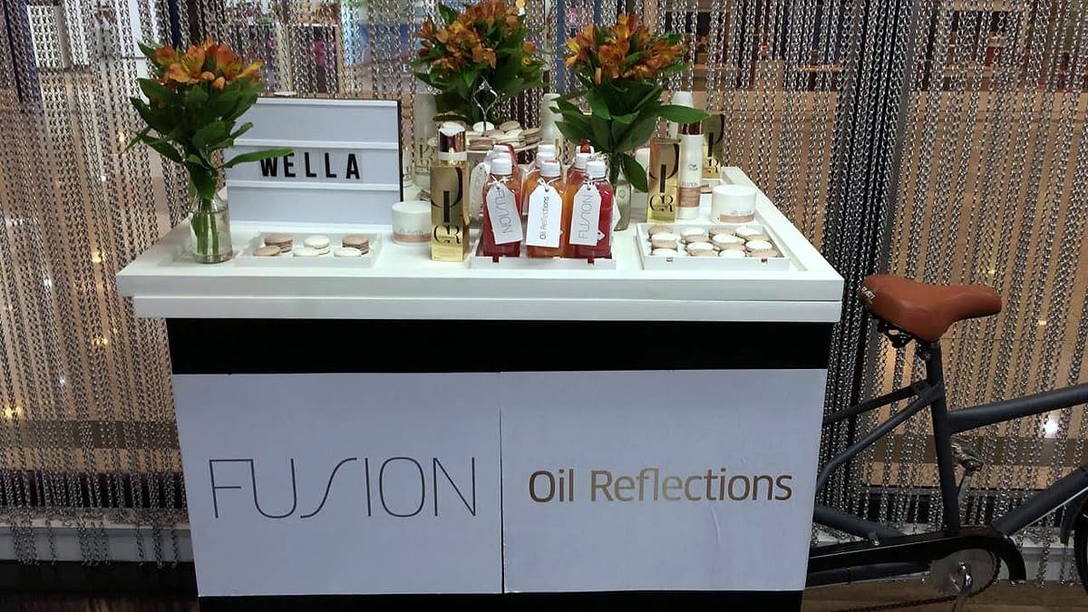 Carrinho branco com painéis 'FUSION' e 'Oil Reflections', letreiro 'WELLA', produtos e flores laranja sobre o tampo.