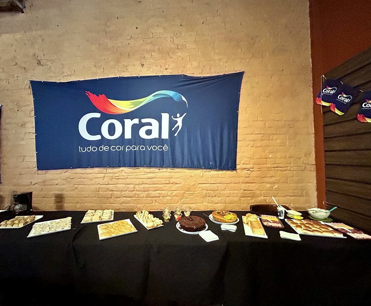 Mesa com toalha preta e bandejas de salgados, bolos e pães em frente a um banner azul da Coral na parede de tijolos.