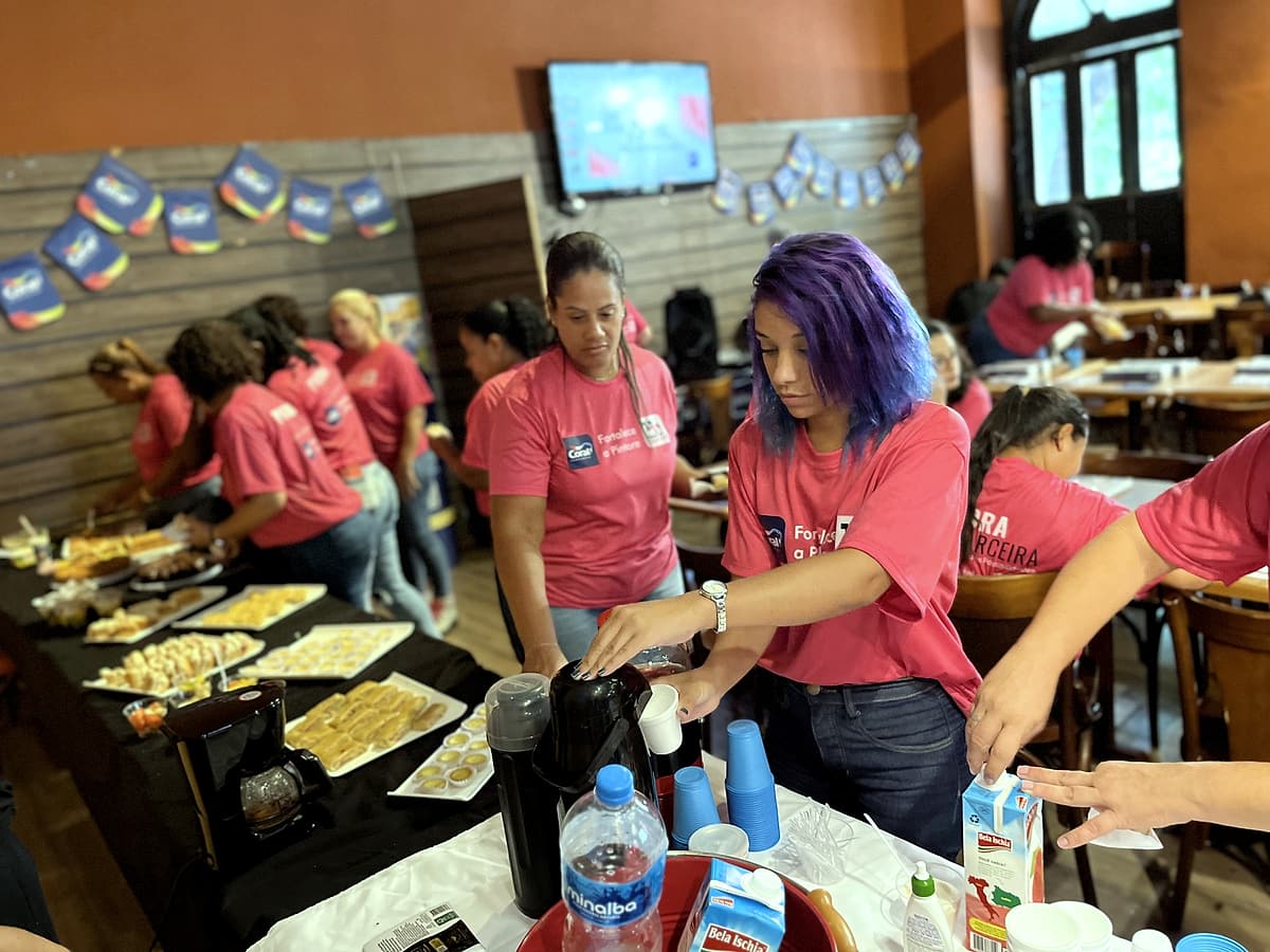 Mulheres com camisetas rosa da Coral se servem de café e salgados em uma mesa de coffee break decorada com bandeirolas da marca.