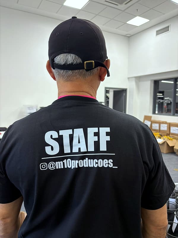 Pessoa de costas vestindo camiseta preta com 'STAFF @m10producoes_' e boné, em sala com caixas de brindes ao fundo.