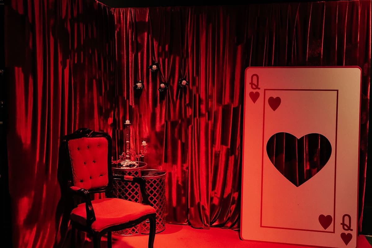 Canto cenográfico com cortinas de veludo vermelho, poltrona capitonê, candelabro e uma carta gigante de Dama de Copas.