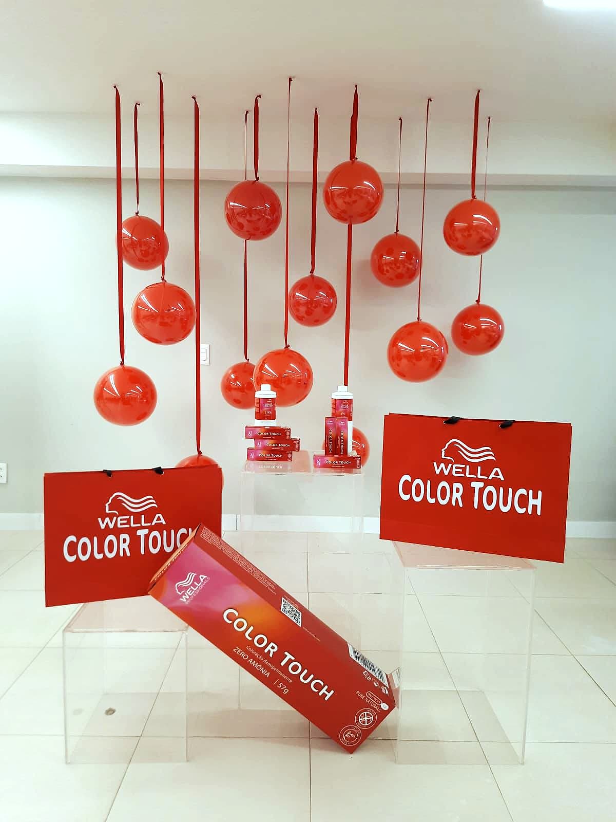 Display com esferas vermelhas suspensas por fitas, cubos de acrílico, sacolas Wella Color Touch e uma embalagem gigante do produto.