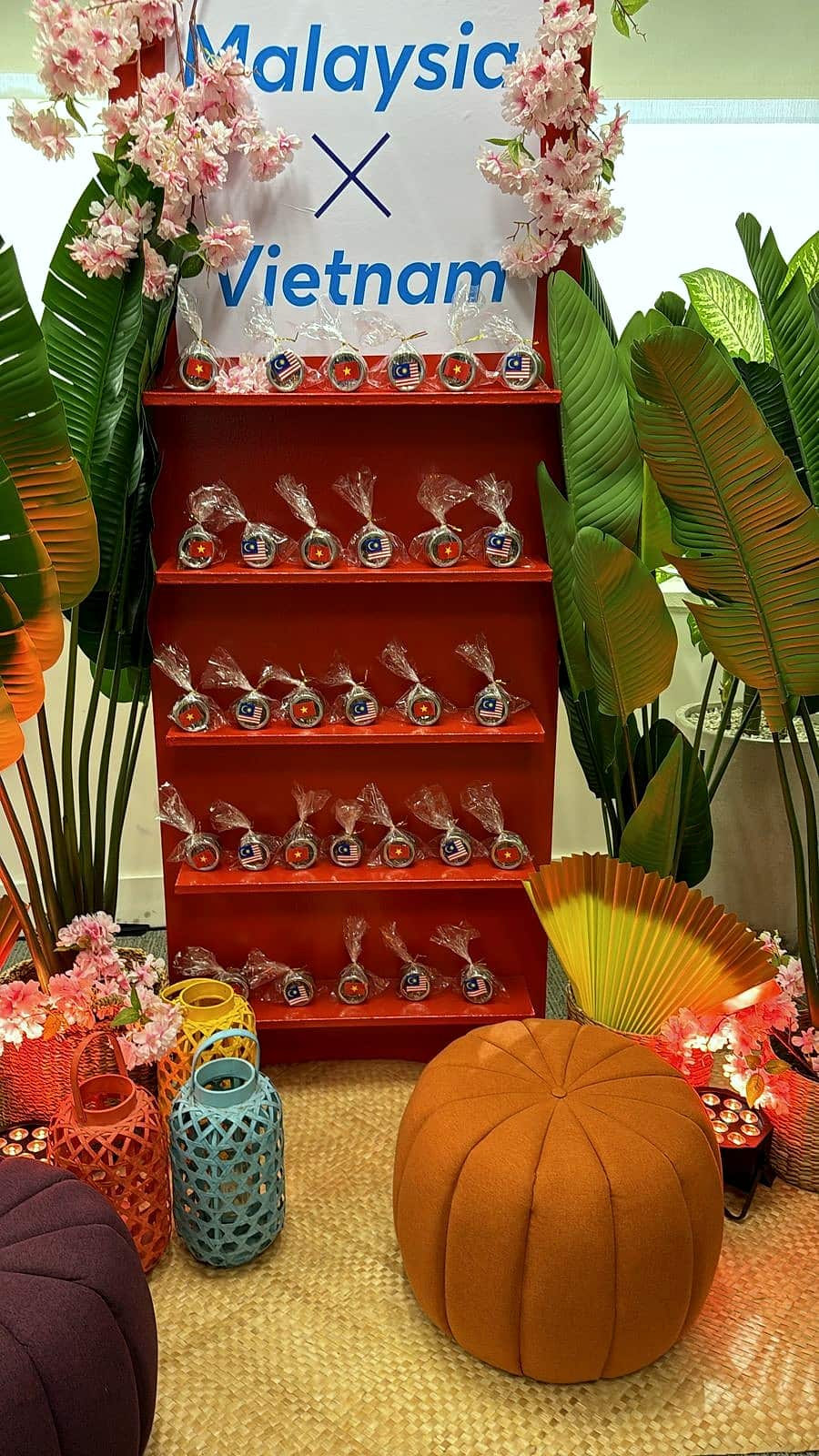 Estante vermelha com lembrancinhas embaladas em plástico, placa com os dizeres Malaysia x Vietnam, flores de cerejeira, folhas tropicais, leque, pufe laranja e lanternas coloridas.