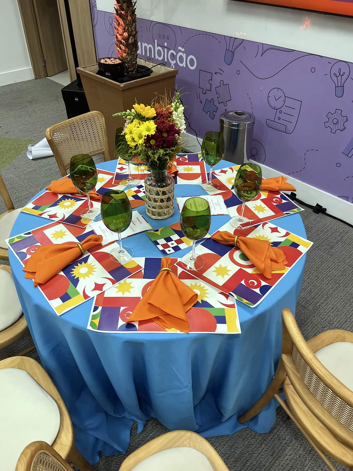 Mesa redonda com toalha azul clara, jogos americanos com estampas de bandeiras, guardanapos laranja, taças verdes e arranjo de flores amarelas e vermelhas.