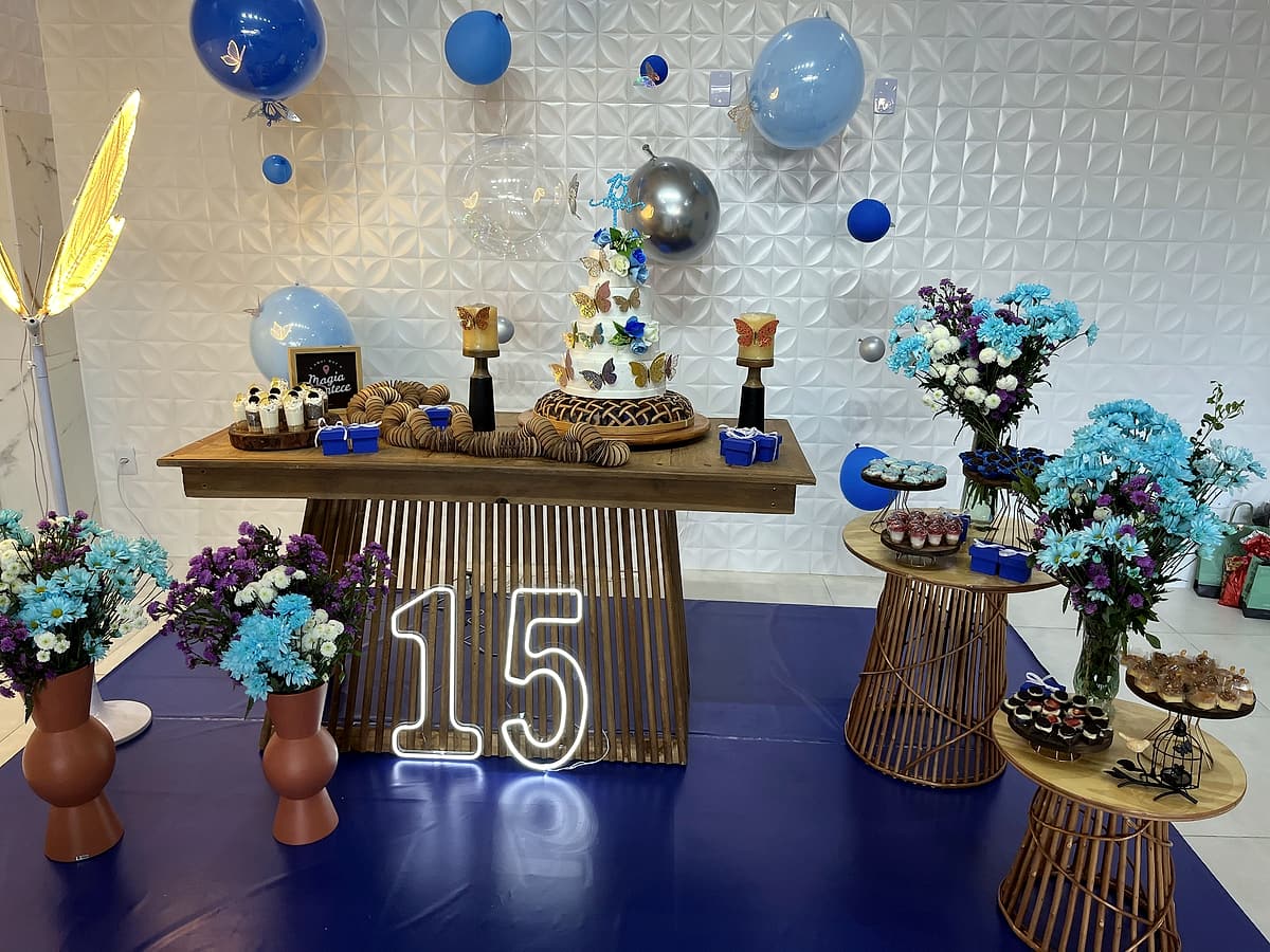 Mesa de madeira com bolo decorado com borboletas, doces, número 15 em neon branco, balões azuis e prateados e vasos com flores azuis e brancas sobre piso azul.