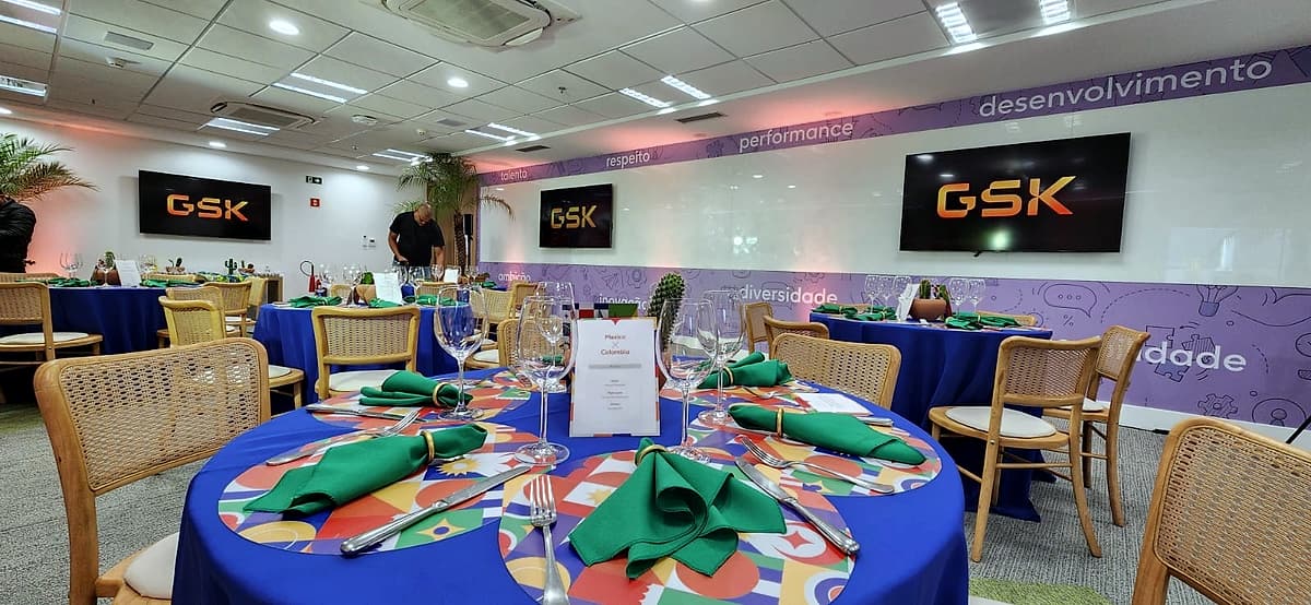 Salão corporativo com mesas redondas de toalha azul, guardanapos verdes e cadeiras de palhinha; telões exibem o logo da GSK.