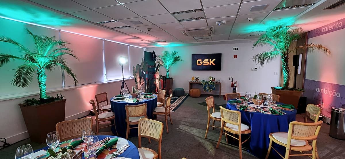 Salão com mesas redondas azuis, palmeiras em vasos e iluminação cênica verde e rosa nas paredes, com logo da GSK no telão.