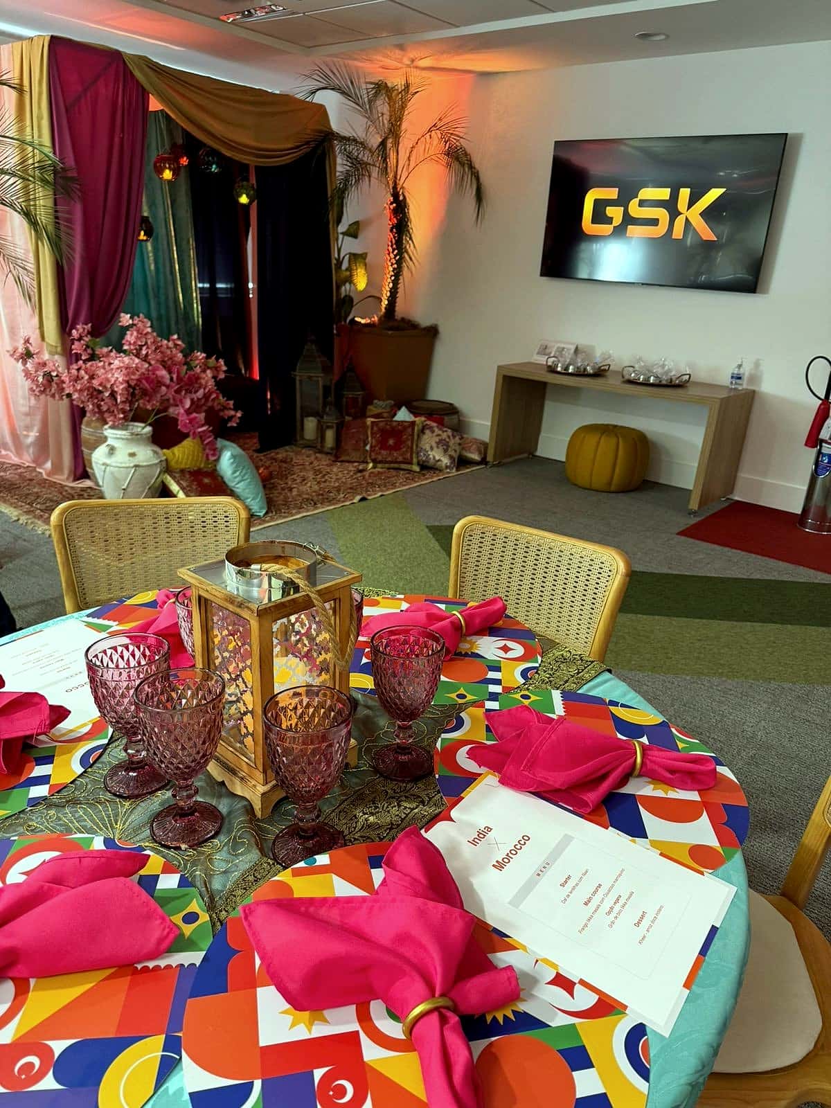 Mesa redonda com toalha estampada colorida, guardanapos cor-de-rosa, taças rosadas, lanterna e cardápio com o título Morocco; ao fundo, lounge com cortinas e telão exibindo o logo da GSK.