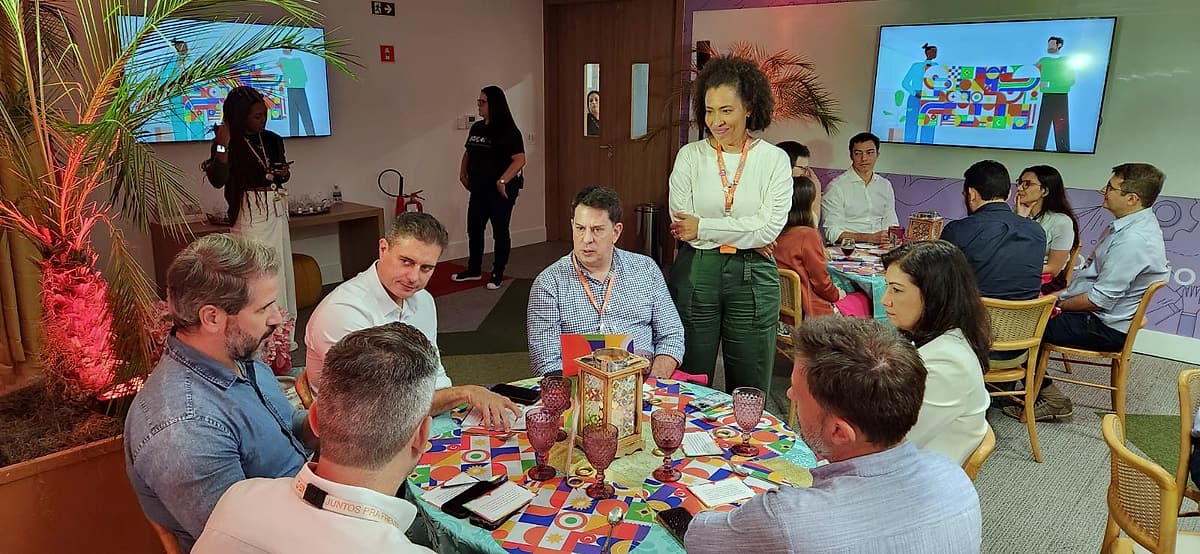 Grupo de convidados reunidos em torno de uma mesa decorada, com uma mulher em pé conversando; ao fundo, telão e plantas com iluminação rosada.