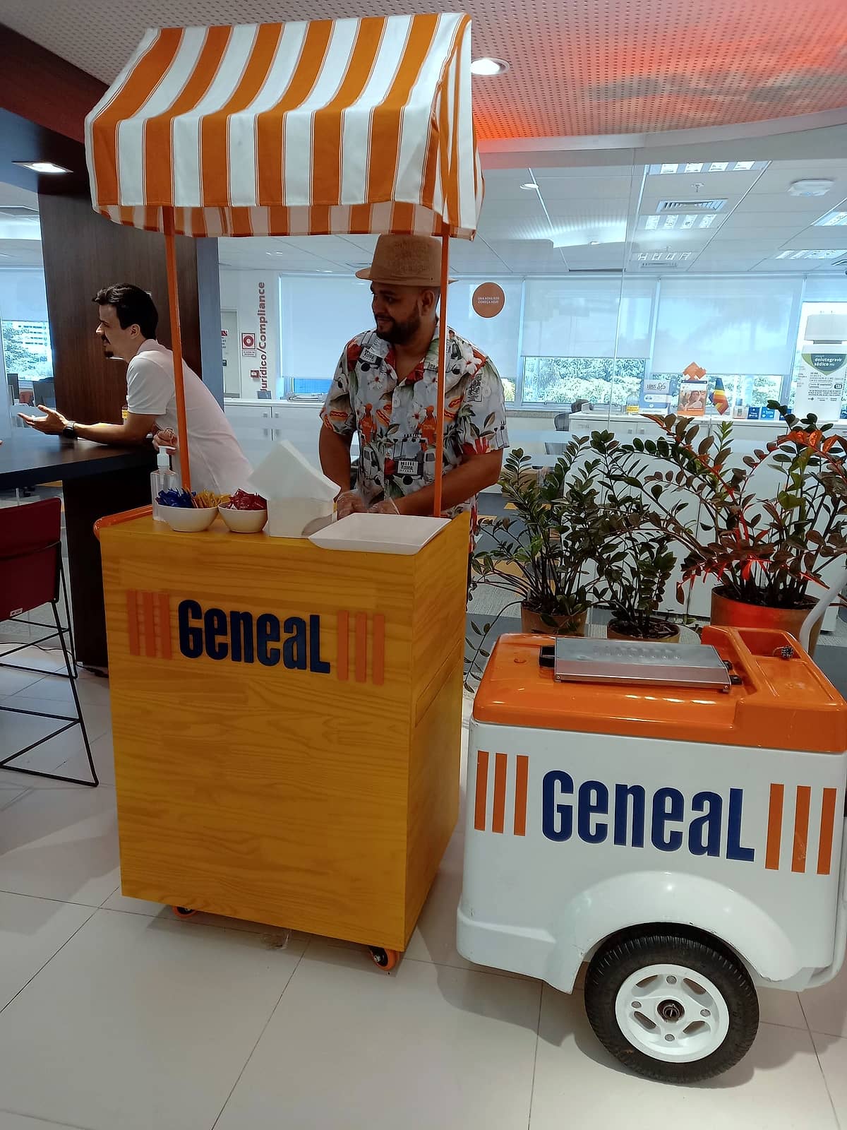 Carrinho de madeira Geneal com toldo listrado laranja e branco e carrinho de sorvete, com atendente de camisa estampada.