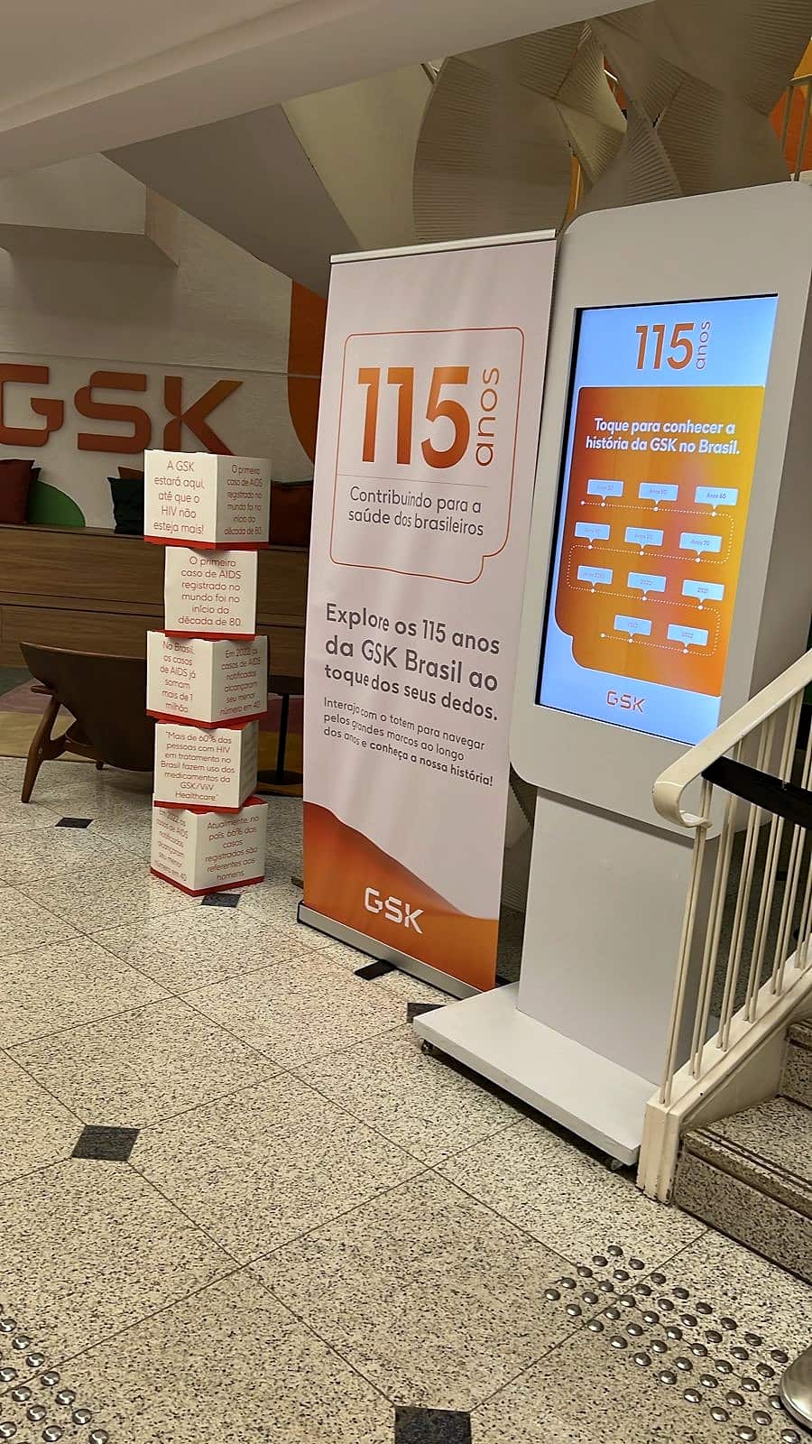 Totem touchscreen com a linha do tempo dos 115 anos da GSK Brasil ao lado de banner 'Explore os 115 anos da GSK Brasil ao toque dos seus dedos'.