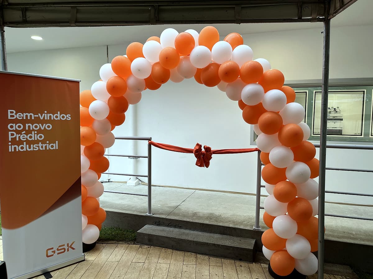 Arco de balões laranja e branco com laço vermelho de inauguração e banner 'Bem-vindos ao novo Prédio industrial'.