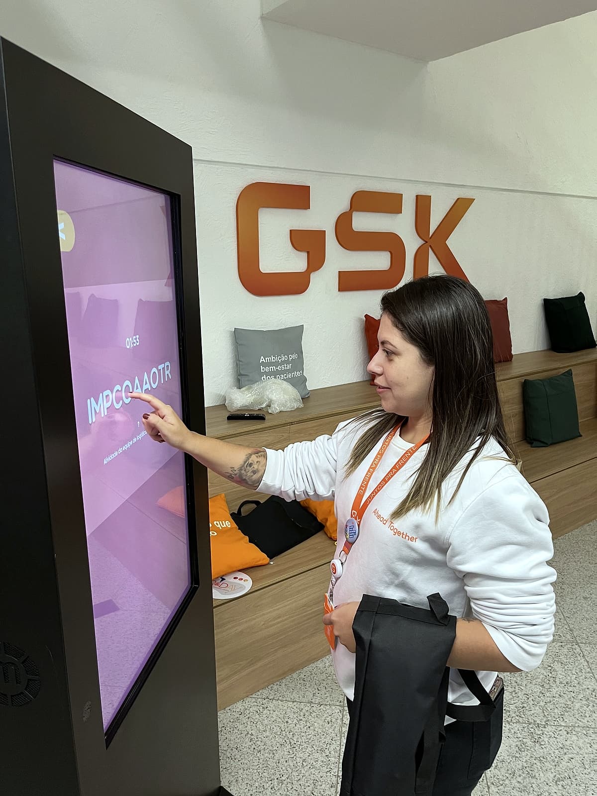 Colaboradora de crachá laranja toca a tela de um totem touch com jogo de palavras, diante do letreiro GSK.