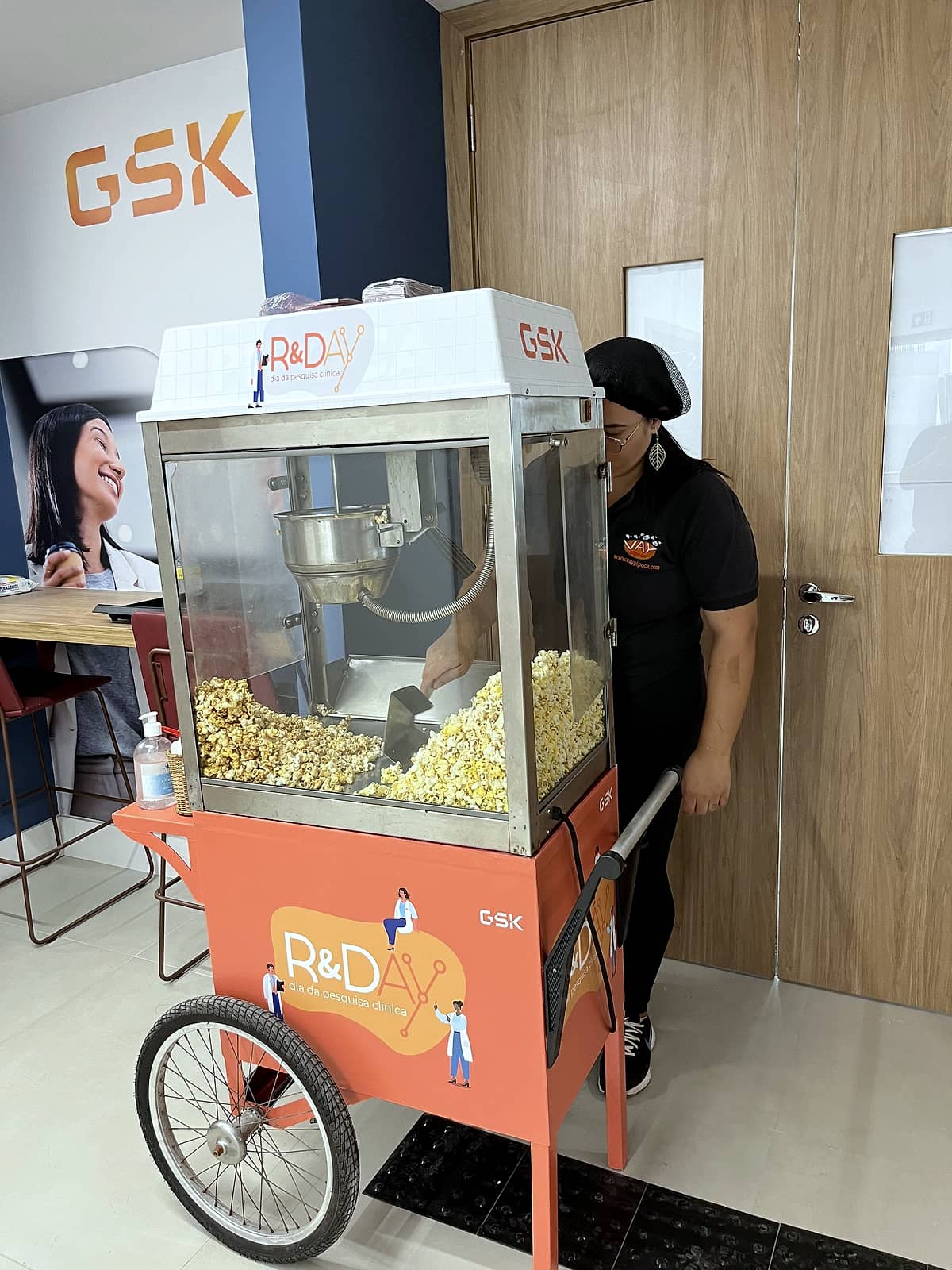 Carrinho de pipoca laranja com adesivo R&Day, dia da pesquisa clínica, e logo GSK, com pipoqueira ao lado.