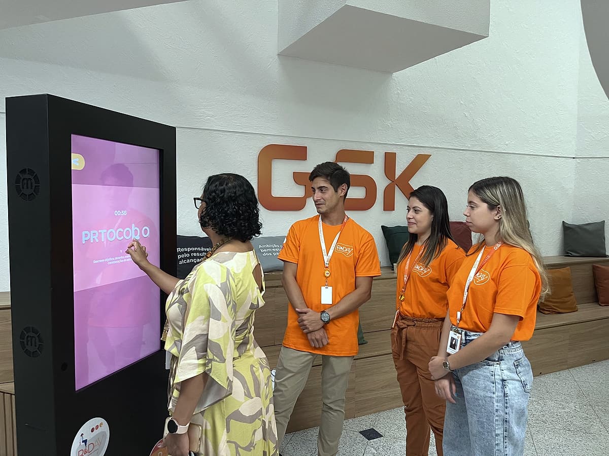 Colaboradora interage com totem touch enquanto equipe de camiseta laranja acompanha, letreiro GSK ao fundo.