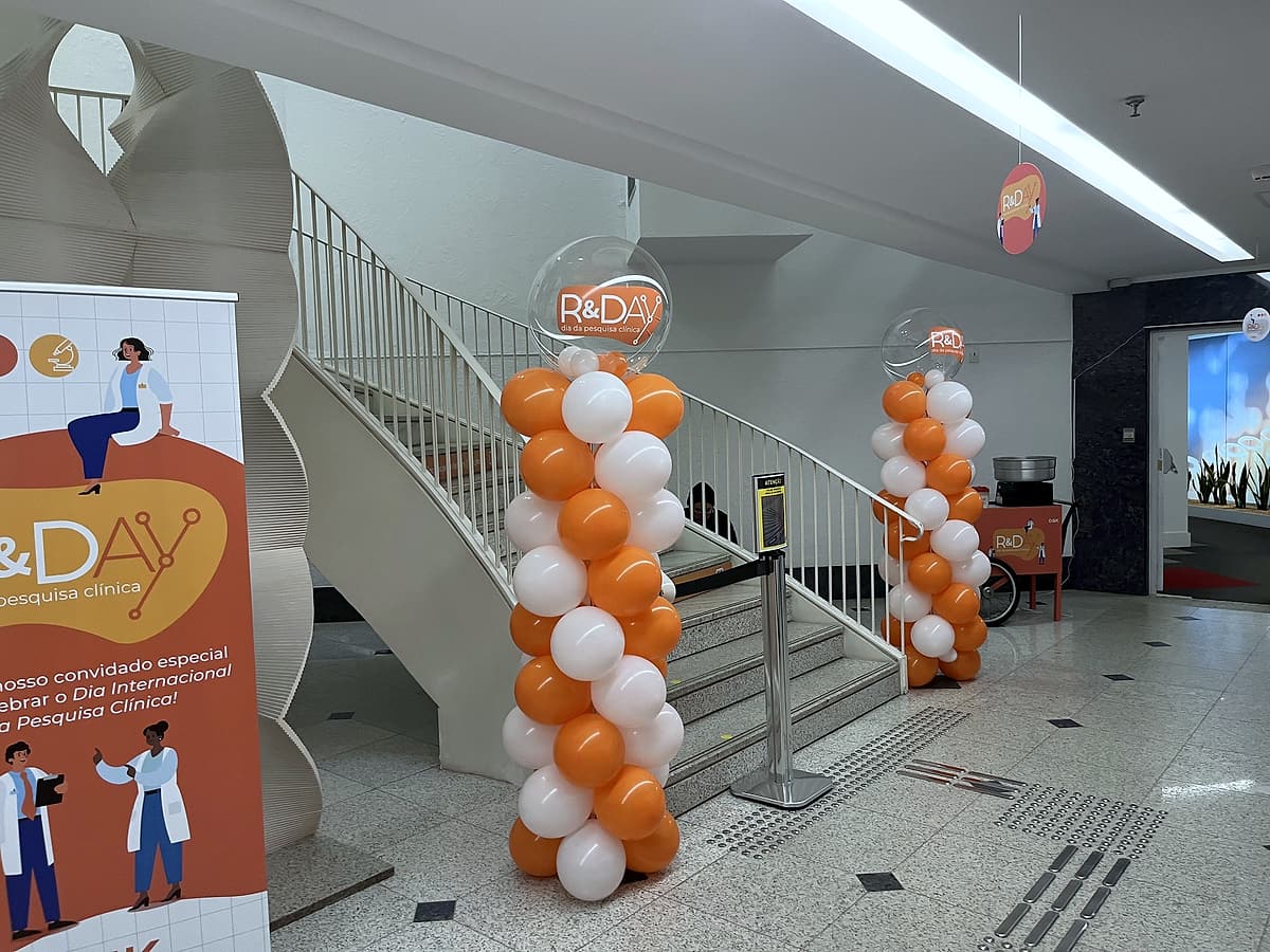 Colunas de balões laranja e brancos com bolha R&Day no hall da escadaria, banner do evento à esquerda.