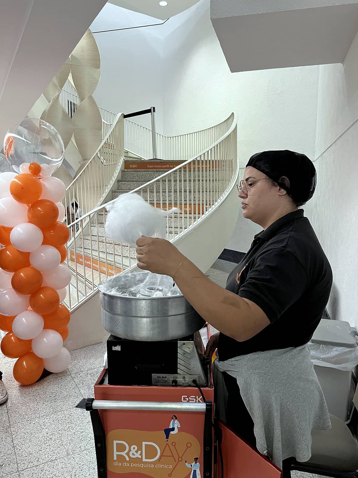 Profissional prepara algodão-doce em carrinho personalizado R&Day ao lado de coluna de balões.