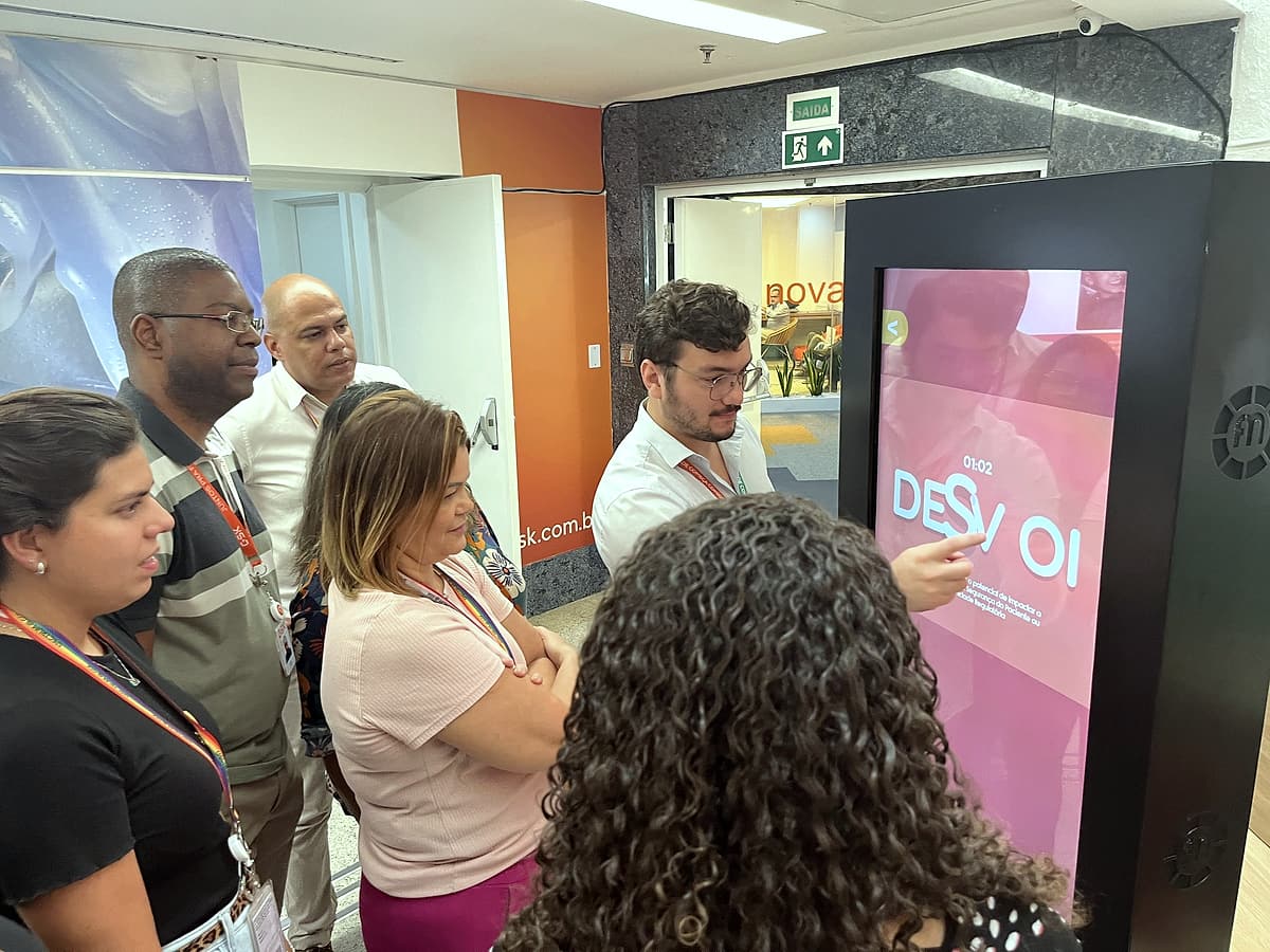 Grupo de colaboradores reunido em volta de um totem touch com um desafio de palavras na tela.