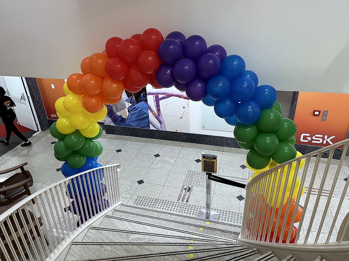Arco de balões nas cores do arco-íris sobre a escadaria, com letreiro GSK ao fundo.