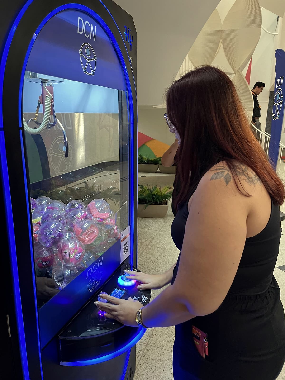 Colaboradora de costas controlando a máquina de garra com luzes azuis e cápsulas de prêmio.