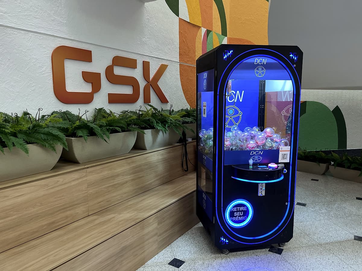 Máquina de garra com luzes azuis e prêmios em cápsulas ao lado do letreiro laranja GSK.