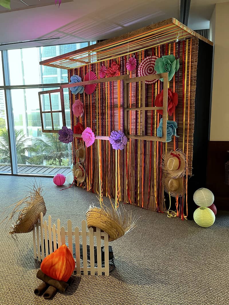 Cenário junino montado com painel de fitas coloridas, janela de madeira, flores de papel, cerquinha e fogueira cenográfica.