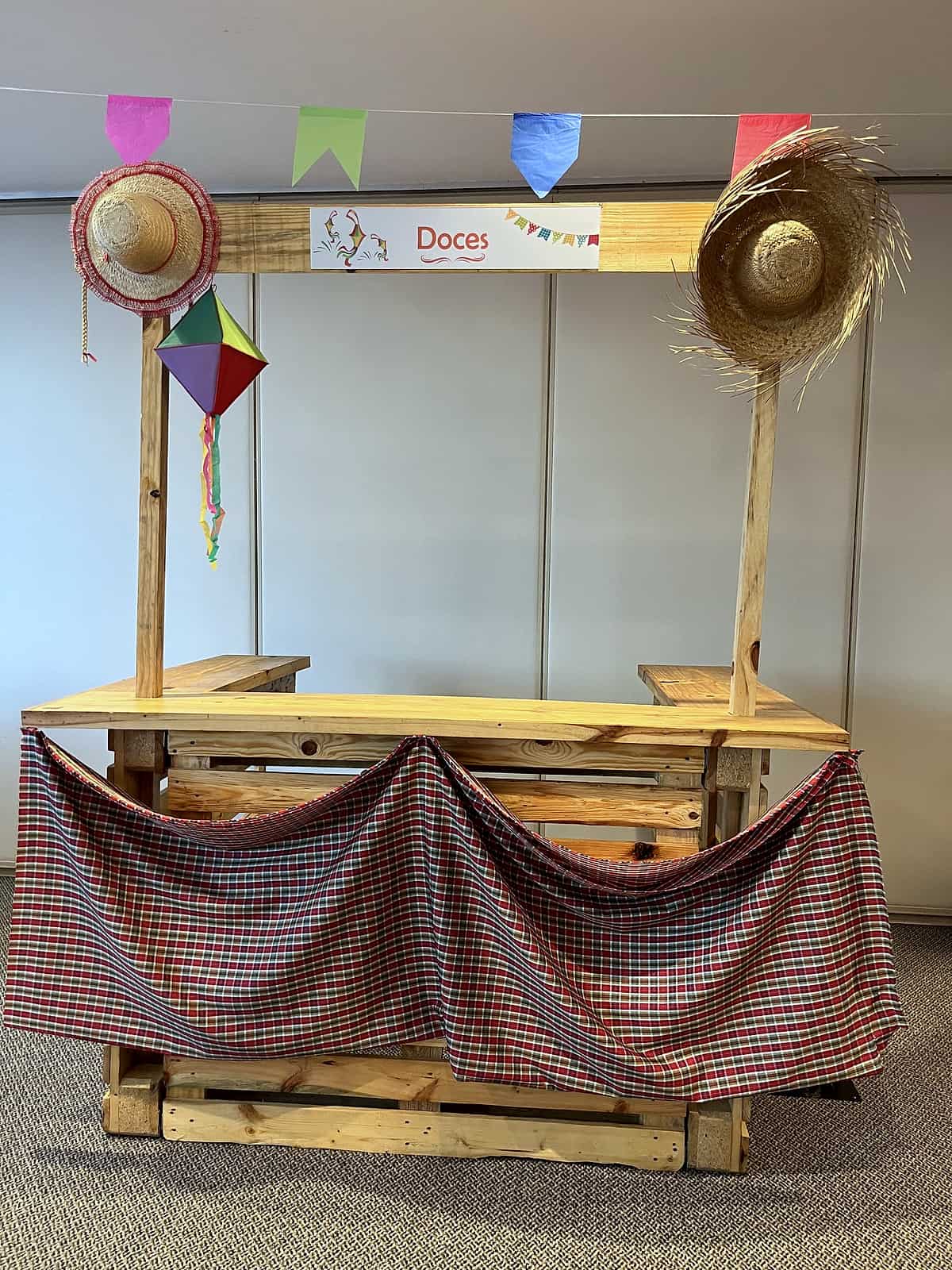 Barraca de madeira de pallets com tecido xadrez, bandeirinhas, balão junino e placa 'Doces'.