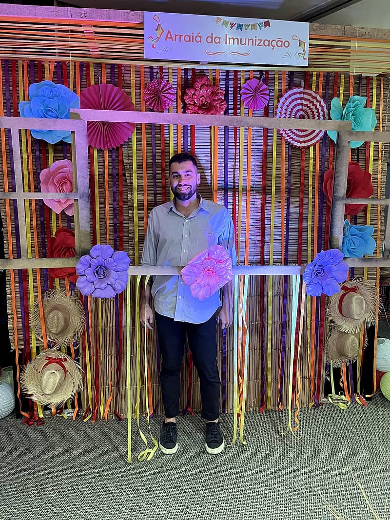 Painel cenográfico junino com fitas coloridas, flores de papel, chapéus de palha e placa 'Arraiá da Imunização', com colaborador posando.