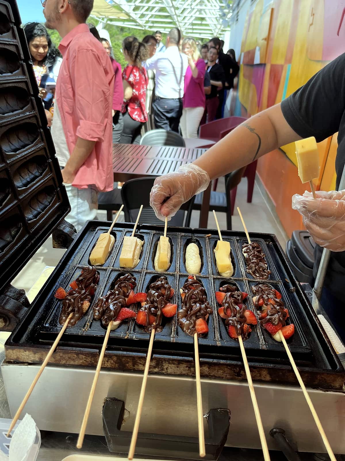 Chapa com espetinhos de massa cobertos de chocolate e morangos, com colaboradores ao fundo.