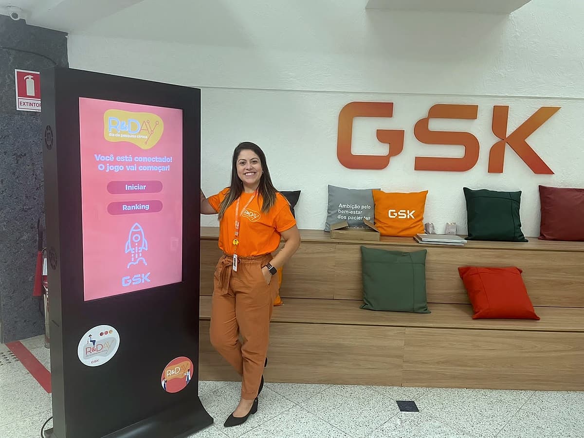 Promotora de camiseta laranja ao lado de totem digital com jogo R&Day, diante do letreiro GSK.
