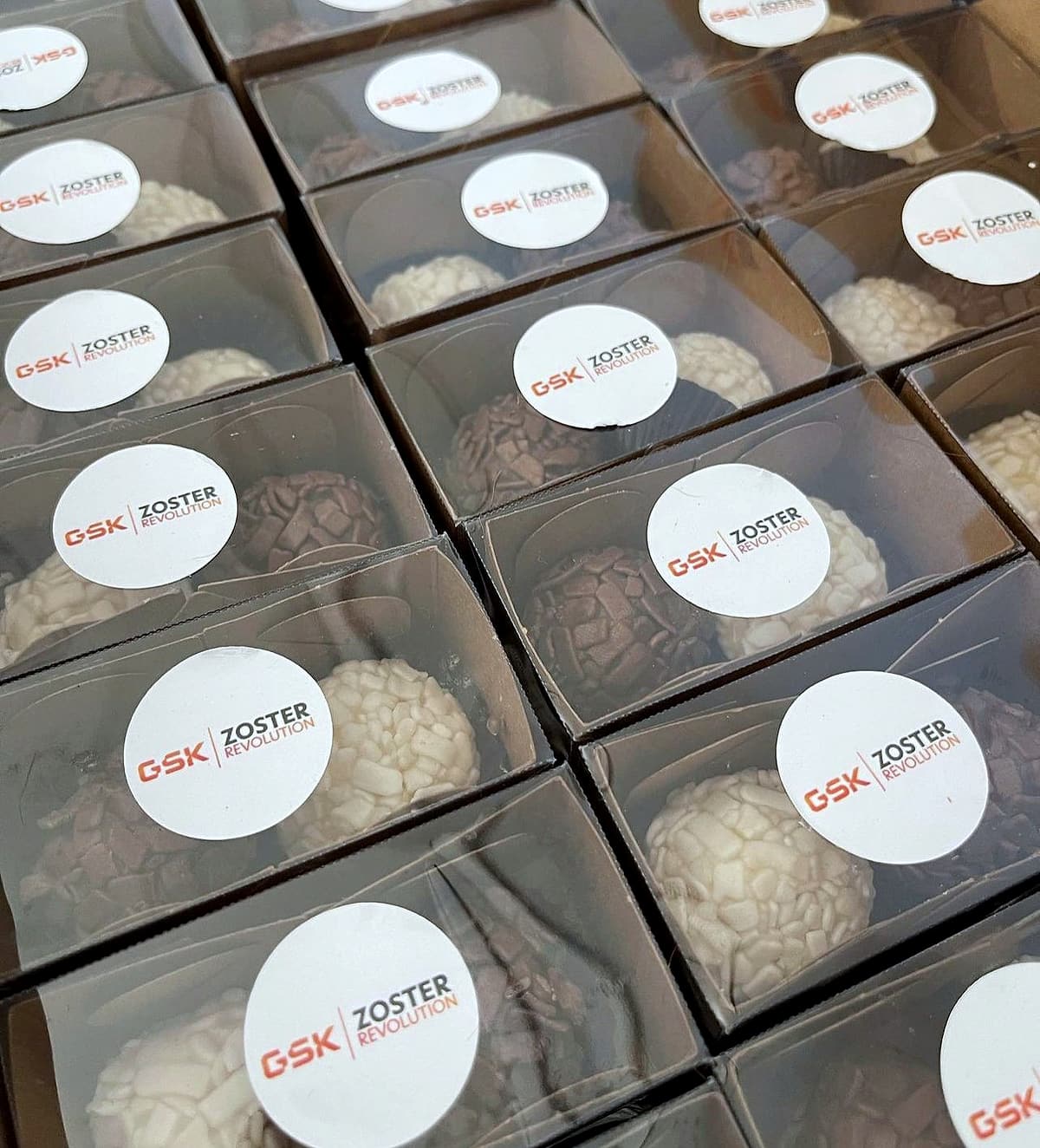 Caixas transparentes com brigadeiros e adesivo redondo GSK Zoster Revolution enfileiradas.